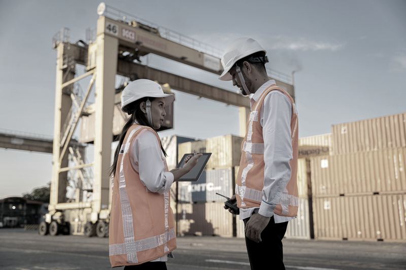 Two terminal workers in hard hats reviewing paperwork beside stacked containers